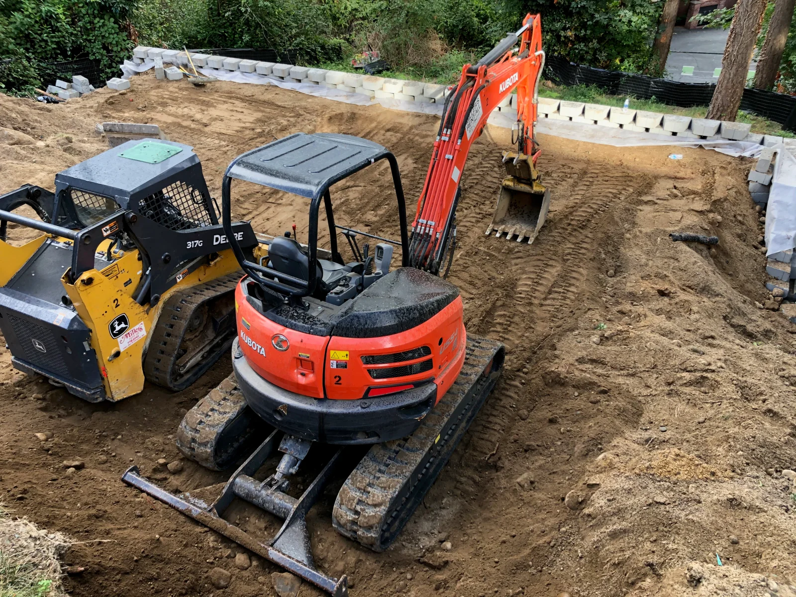 Excavation equipment grading soil with concrete retaining wall blocks staged on prepared site